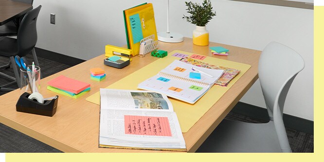 Back-to-school desk setup featuring Post-it® Super Sticky Notes and flags, a yellow Post-it display board, and annotated textbook and notebook on a wood desk.
