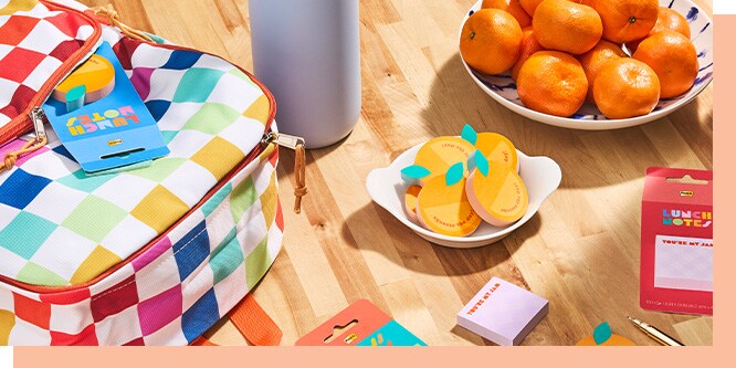 Post-it Brand Lunch Notes in clementine, blue, and pink designs displayed in a back-to-school flat-lay scene with a colorful checkered lunch bag and fresh fruit.

