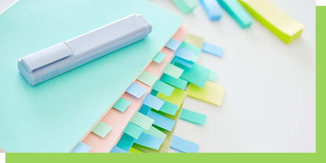 Pastel notebooks with colorful sticky tabs and a gray pen arranged neatly on a white surface.
